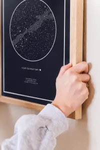 A hand hanging a wood framed navy star map print on a wall, captioned “I love you Milky Way”